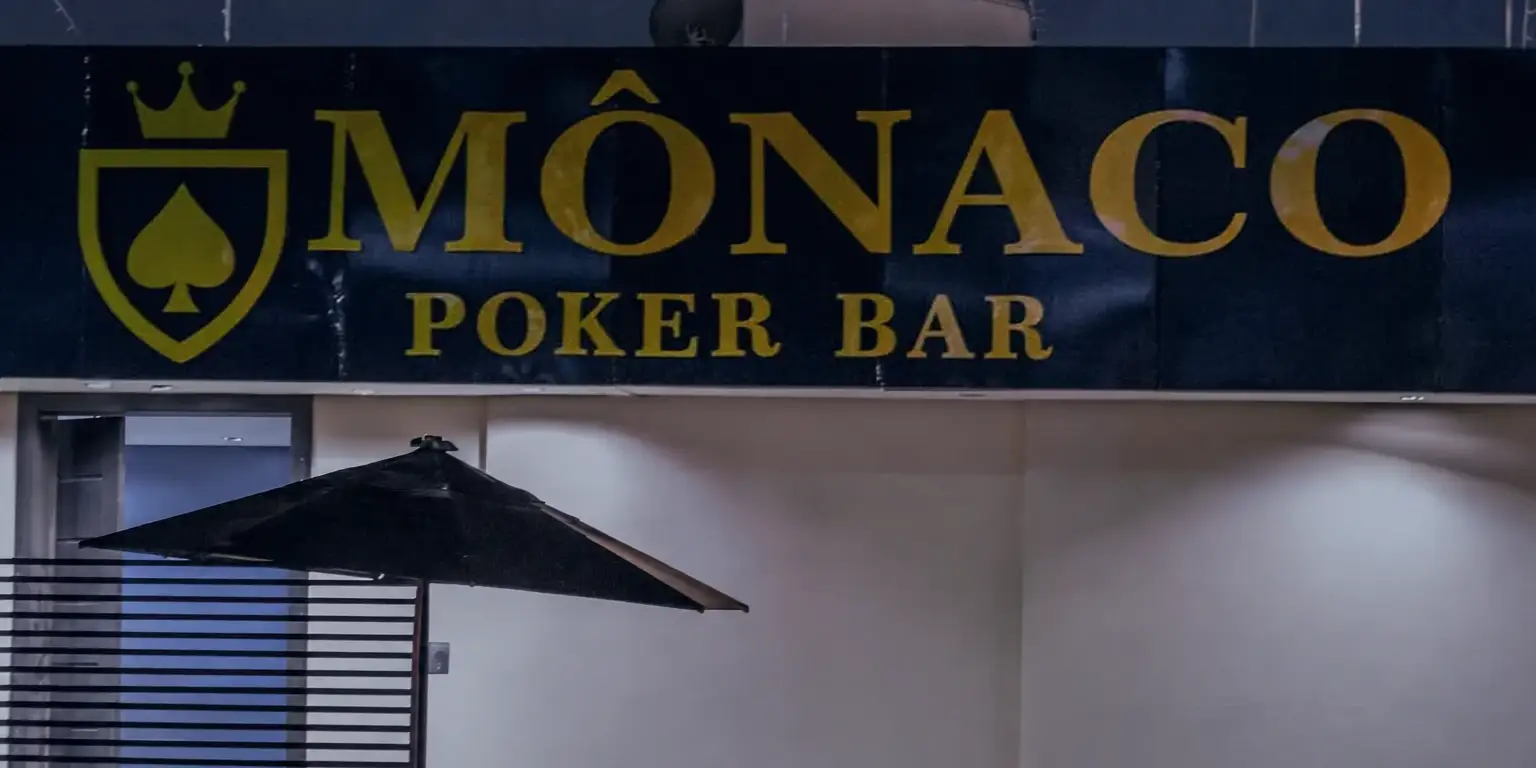 A entrada do Monaco à noite: o letreiro azul e dourado com o escudo, a parede iluminada, o guarda-sol e a porta de ripas, na Rua dos Alecrins, 365