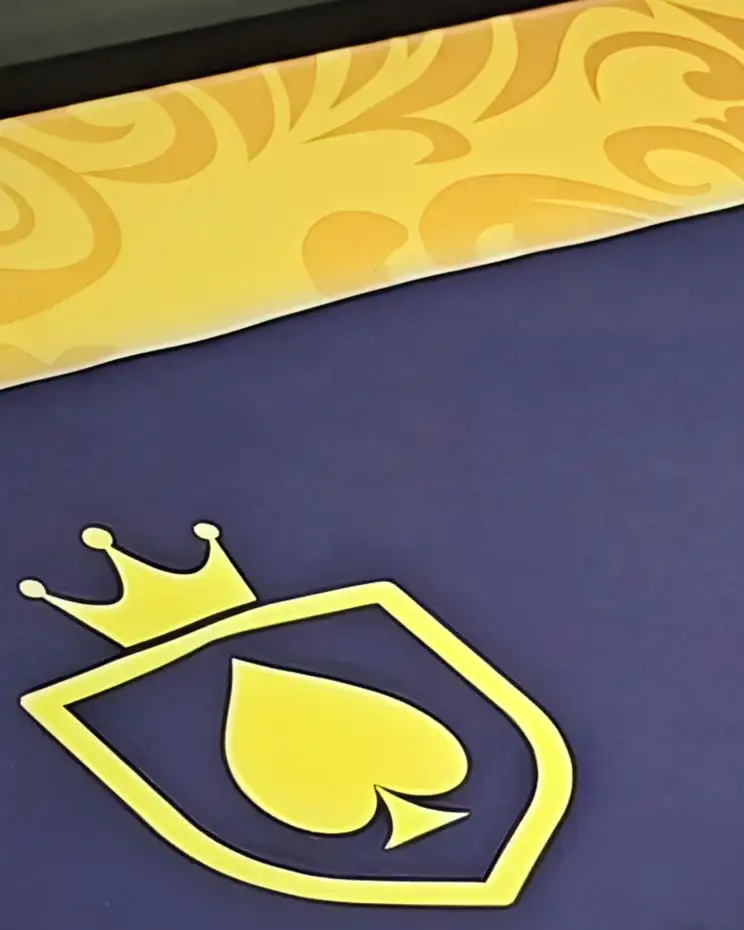 O feltro azul de uma mesa do Monaco, de perto: o escudo dourado da casa, com a coroa, e o trilho com arabescos na borda